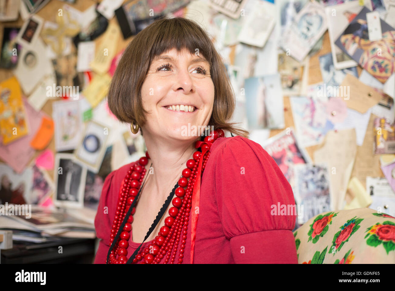 A lady in red blouse and necklaces smiling with wall of photos and cards in the background. Stock Photo