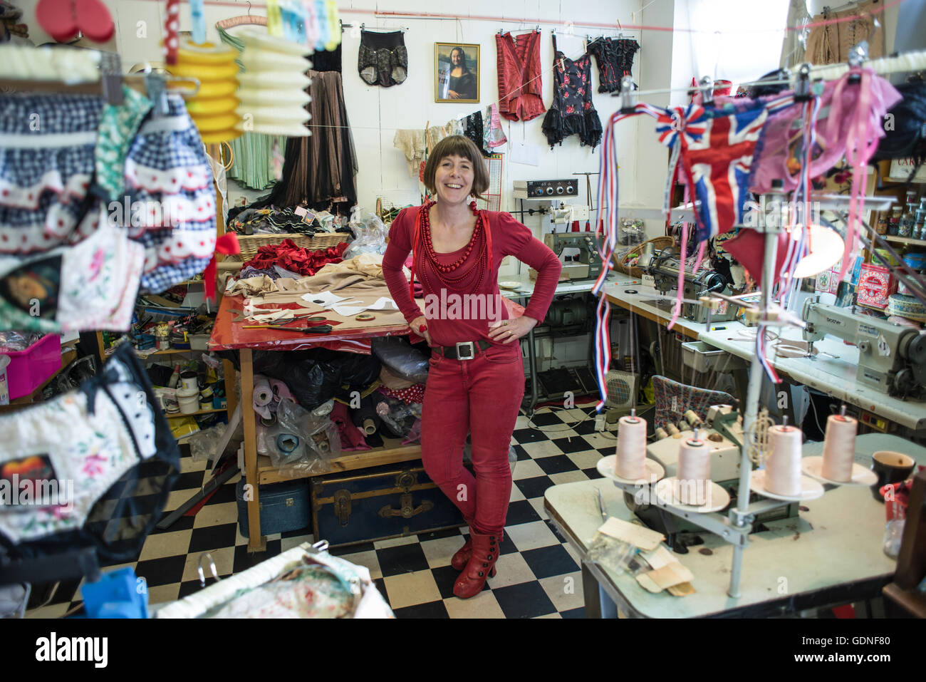A woman in red blouse, trousers, and necklaces smiling and standing in the middle of a tailoring shop. Stock Photo