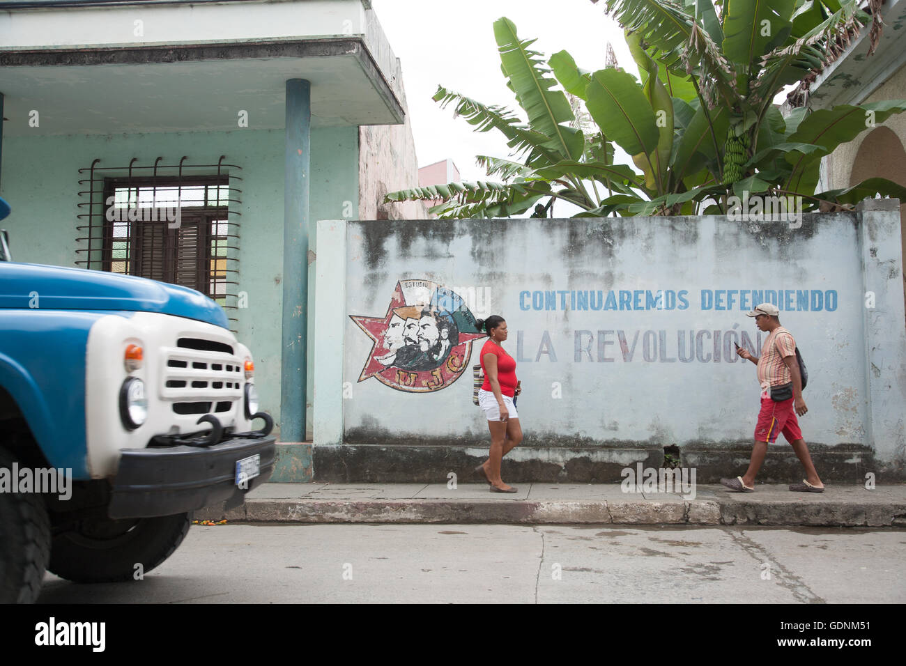 People walking past a pro-revolution mural, one checking his mobile phone, in Baracoa, eastern Cuba (June 2016) Stock Photo