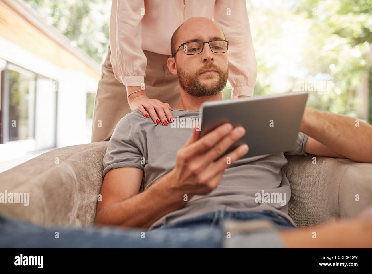 Indoor shot of a mature man sitting on sofa using digital tablet, with woman standing behind him. Couple at home in living room. Stock Photo