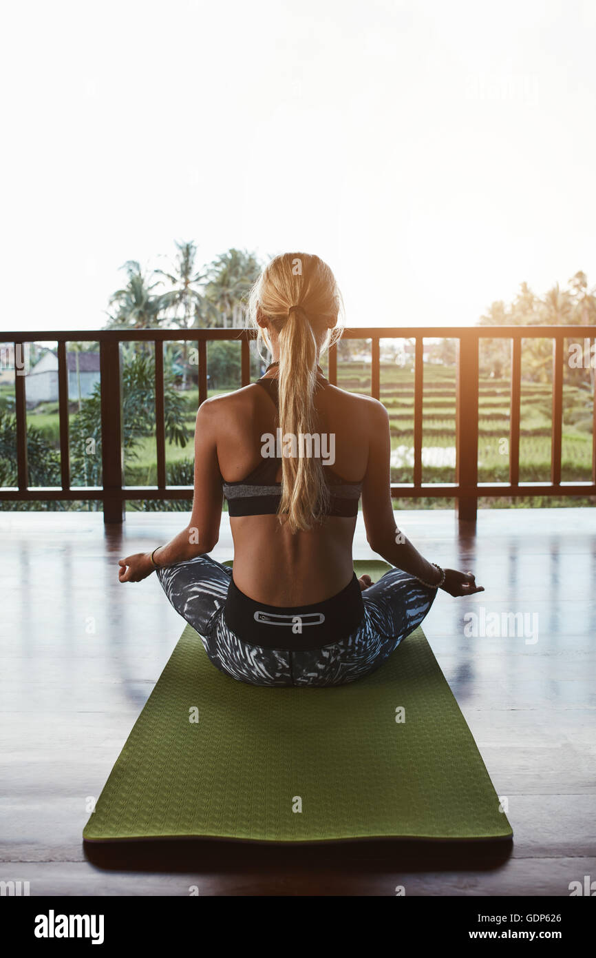 Rear view shot of young woman sitting in lotus yoga pose on exercise mat. Fitness female model doing meditation at health club. Stock Photo