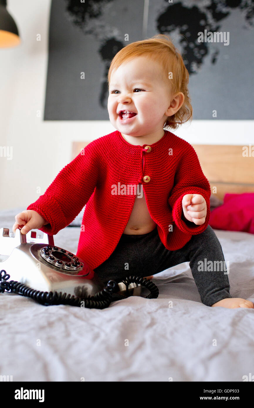 Baby girl sitting on bed playing with landline telephone Stock Photo
