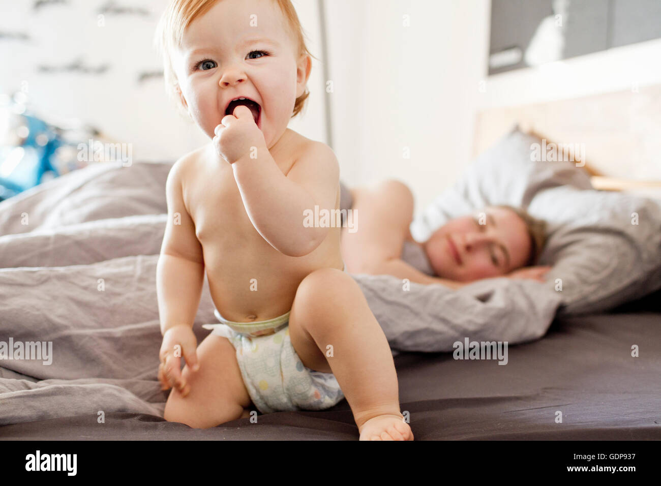 Baby girl sitting on bed with tired mother Stock Photo