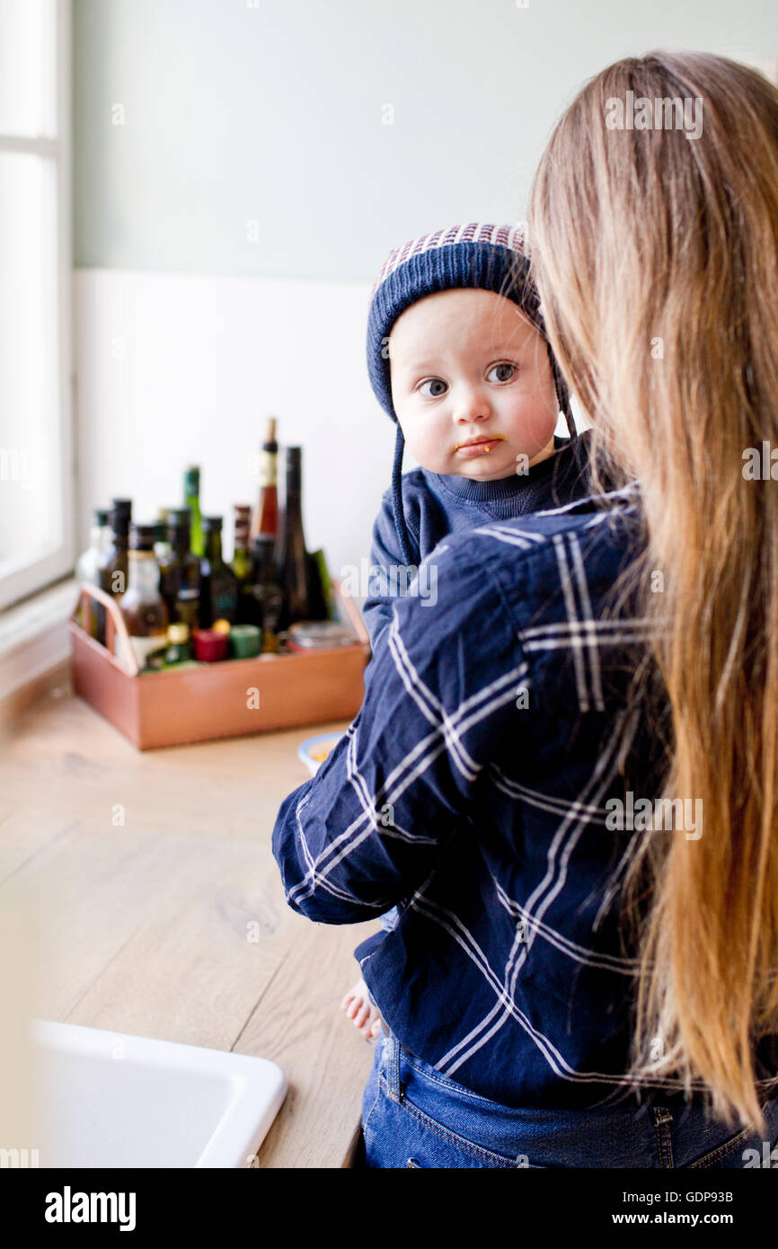 Woman carrying baby son wearing knit hat in kitchen Stock Photo