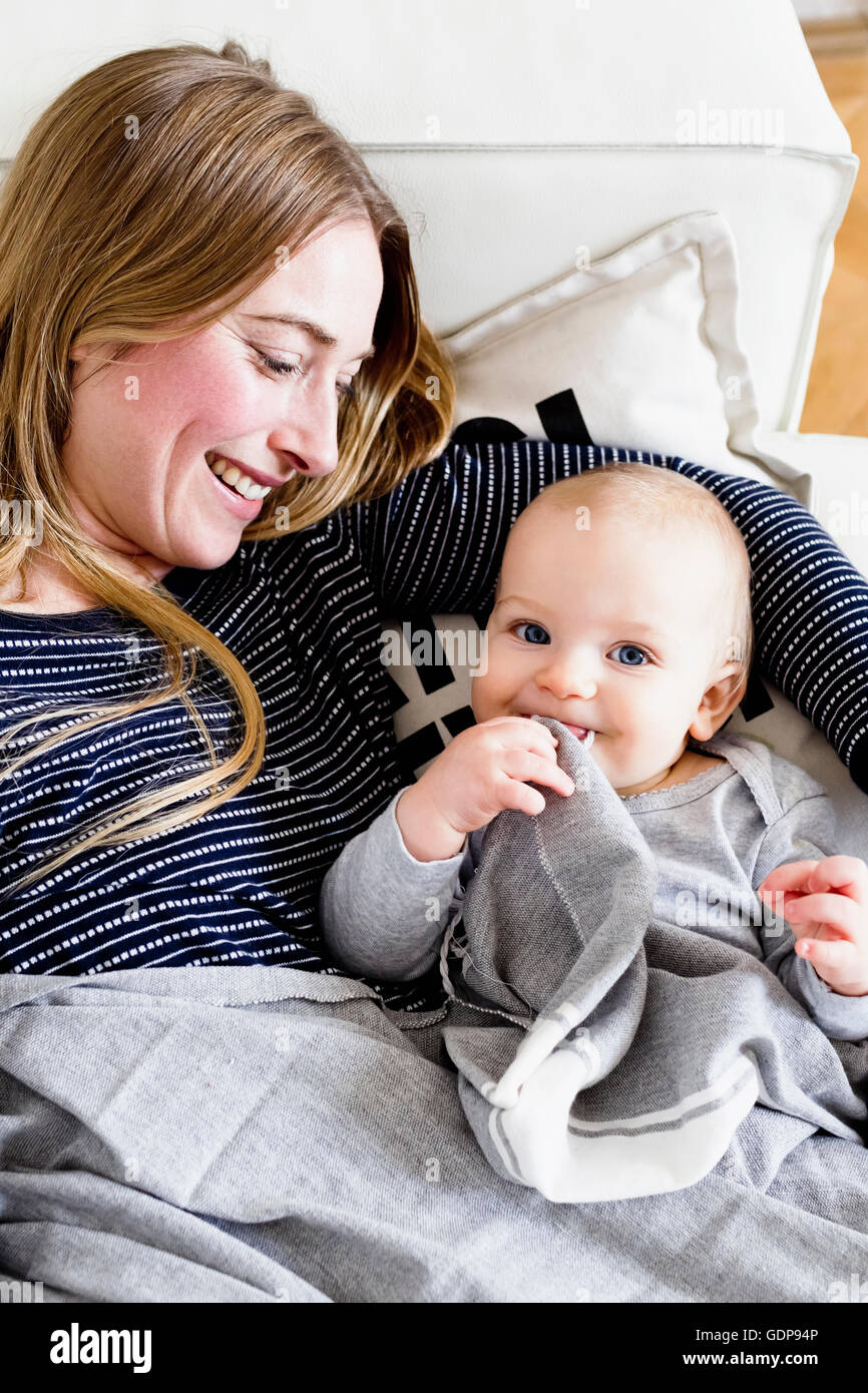 Baby girl reclining on sofa with mother sucking comfort blanket Stock Photo