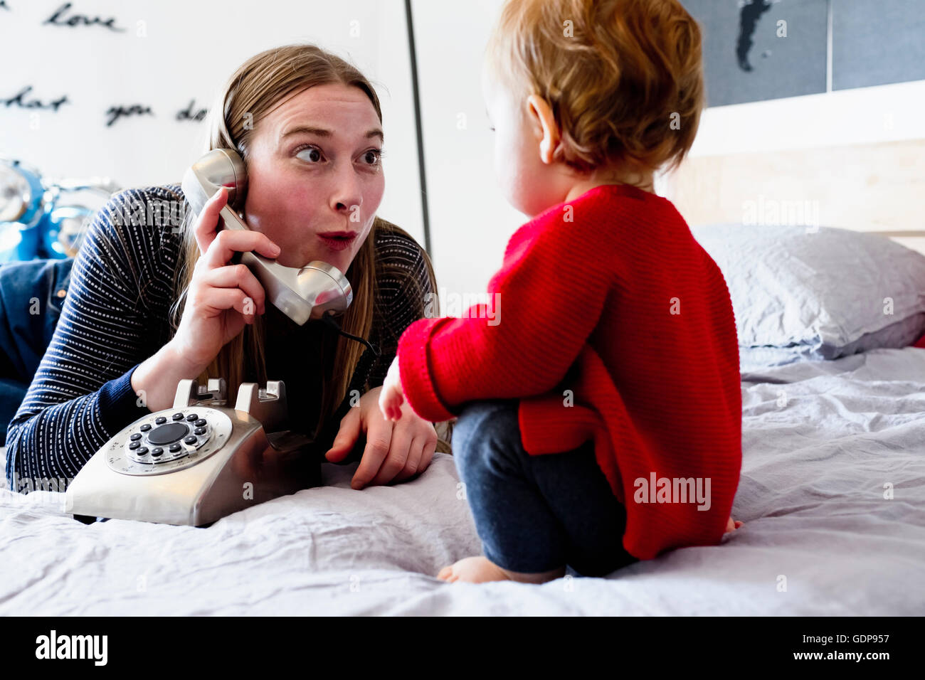 Baby girl and mother on bed pulling face at landline telephone Stock
