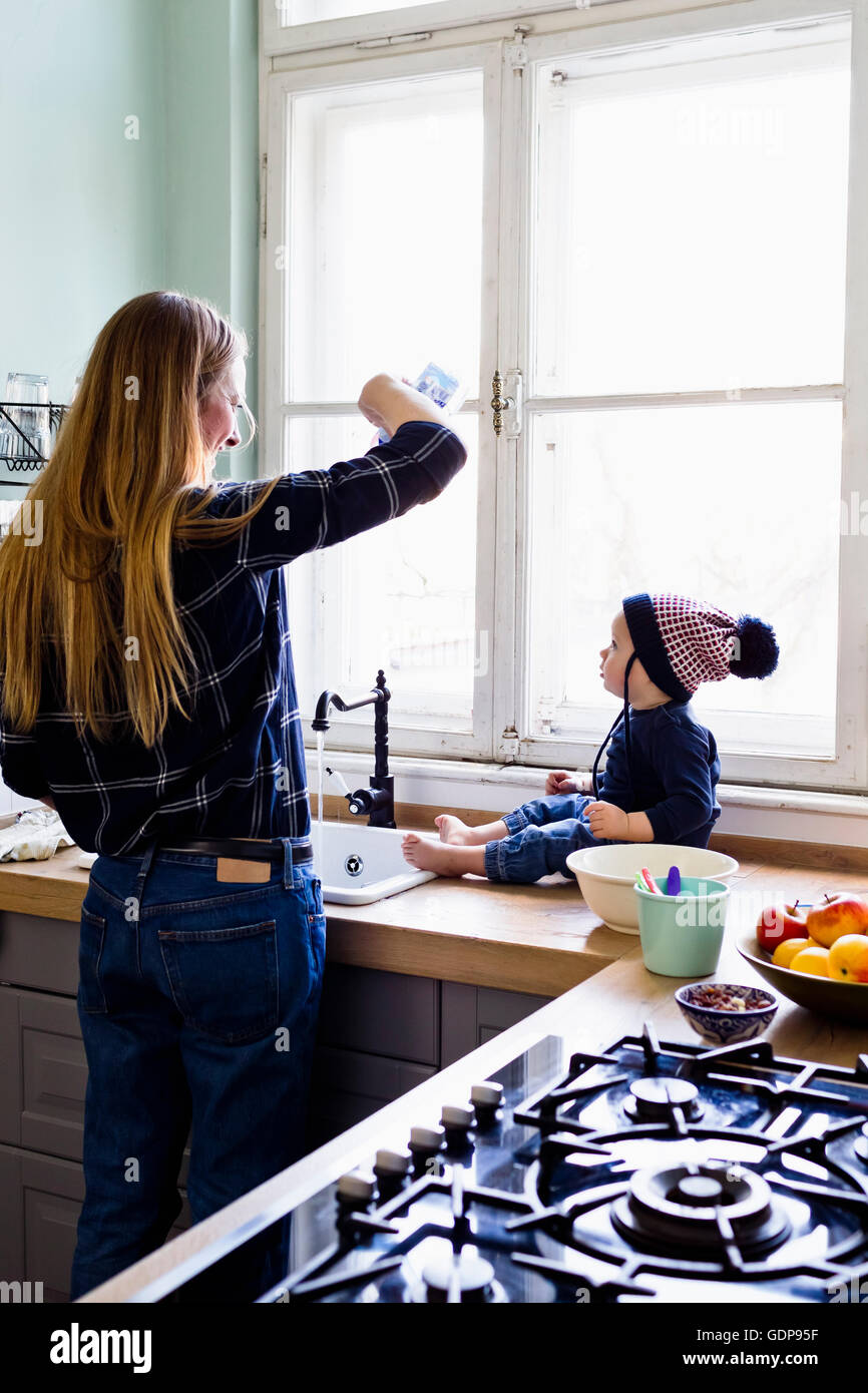 Mid adult woman pouring water into kitchen sink for baby son Stock Photo