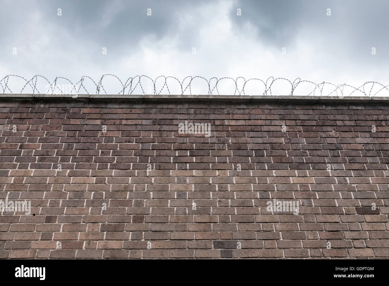 Looking up at a high brick wall with barbed wire on top and dark clouds above Stock Photo