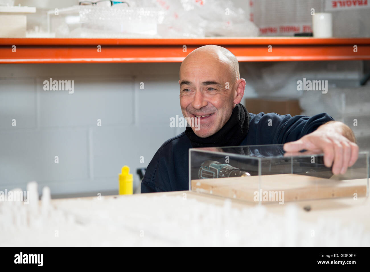 A smiling caucasian man holding a transparent acrylic case in place while drilling holes on its sides Stock Photo