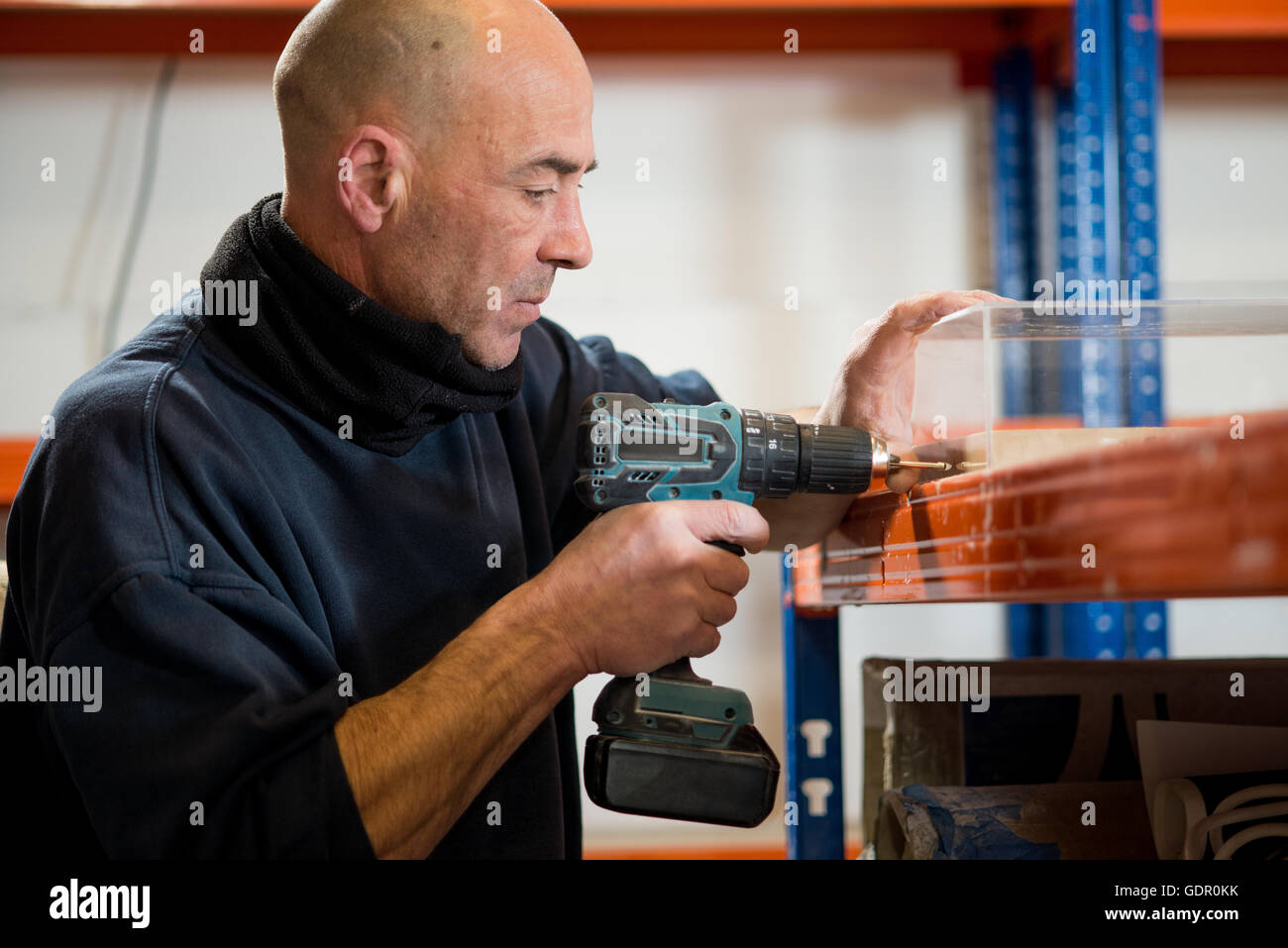 A man in a dark jacket drilling holes on one side of a transparent acrylic case using a cordless drill. Stock Photo