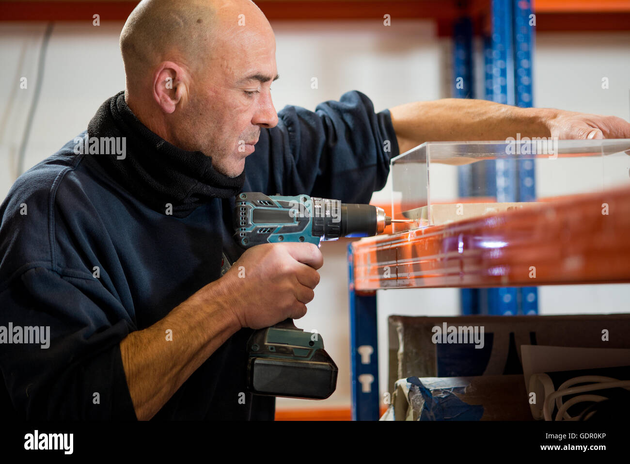 A man in a dark jacket drilling holes on one side of a transparent acrylic case using a cordless power drill. Stock Photo