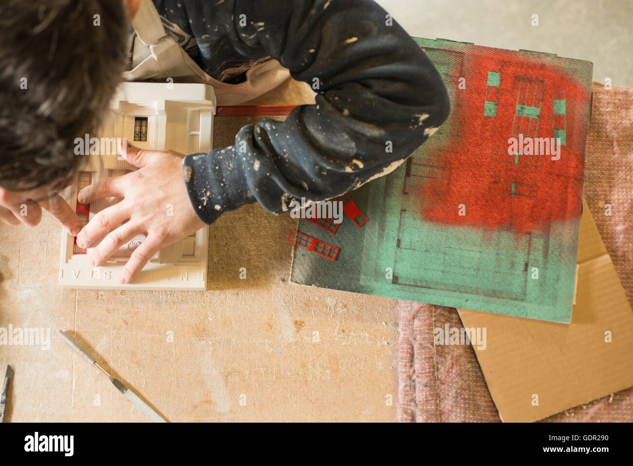 Overhead of a man placing miniature red painted windows on a plaster scale model building on a wooden table. Stock Photo