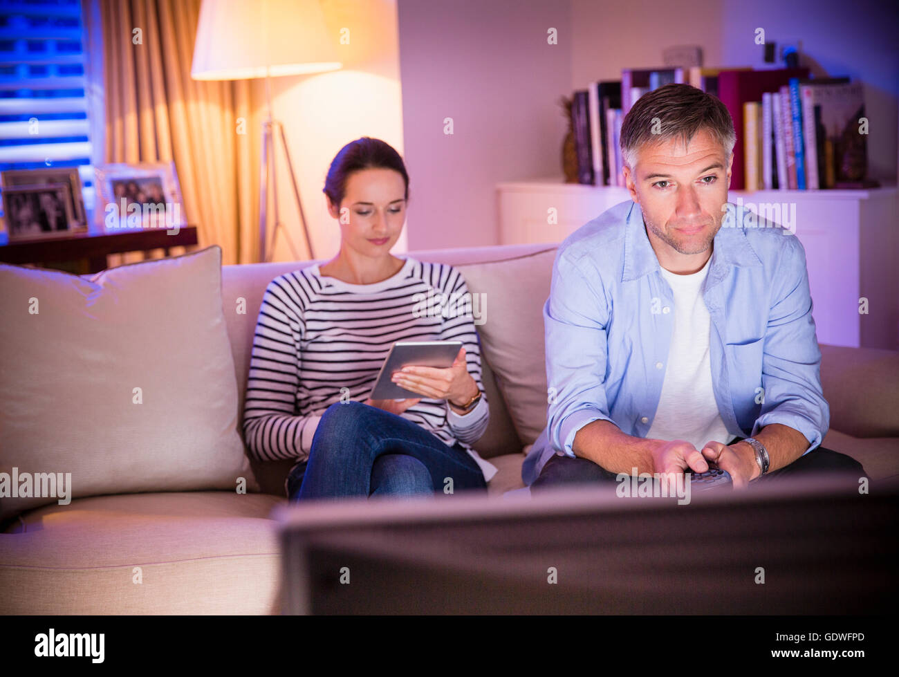 Wife using digital tablet next to husband watching TV in living room Stock Photo