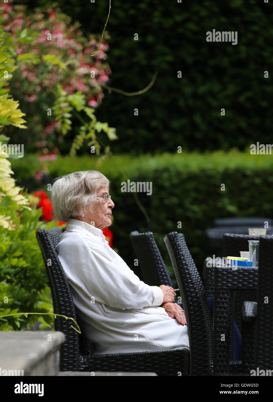 Elderly lady sitting alone and smoking outside in a garden on a rattan chair.Wearing glasses and looking deep in thought. Stock Photo