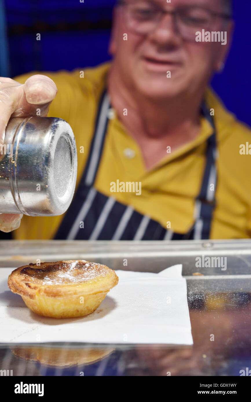 Man sprinkling icing sugar on Pastel de Nata custard tart at a food stall during festival UK Stock Photo