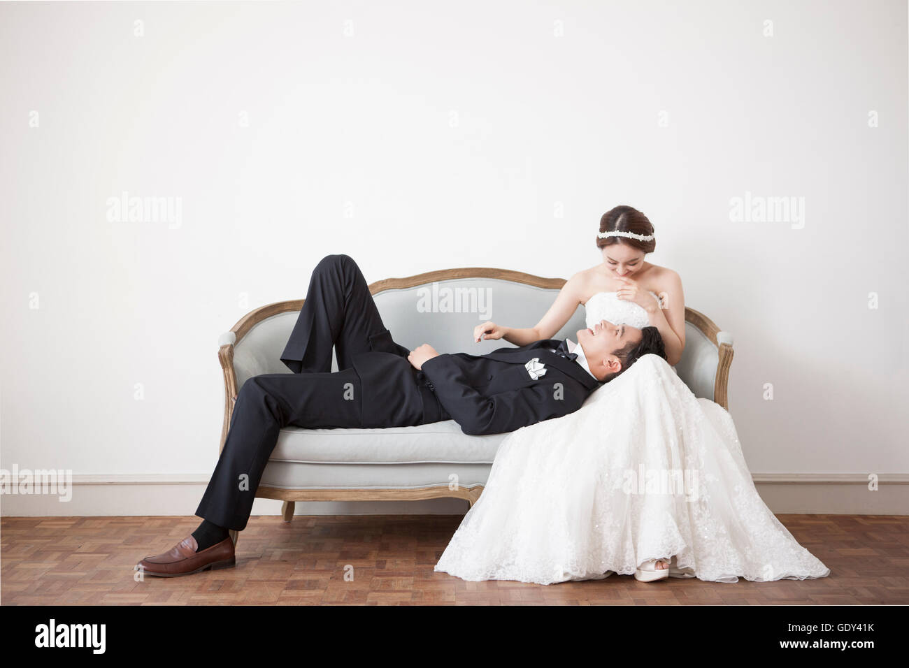 Smiling bride sitting on sofa and groom lying down on her lap Stock Photo