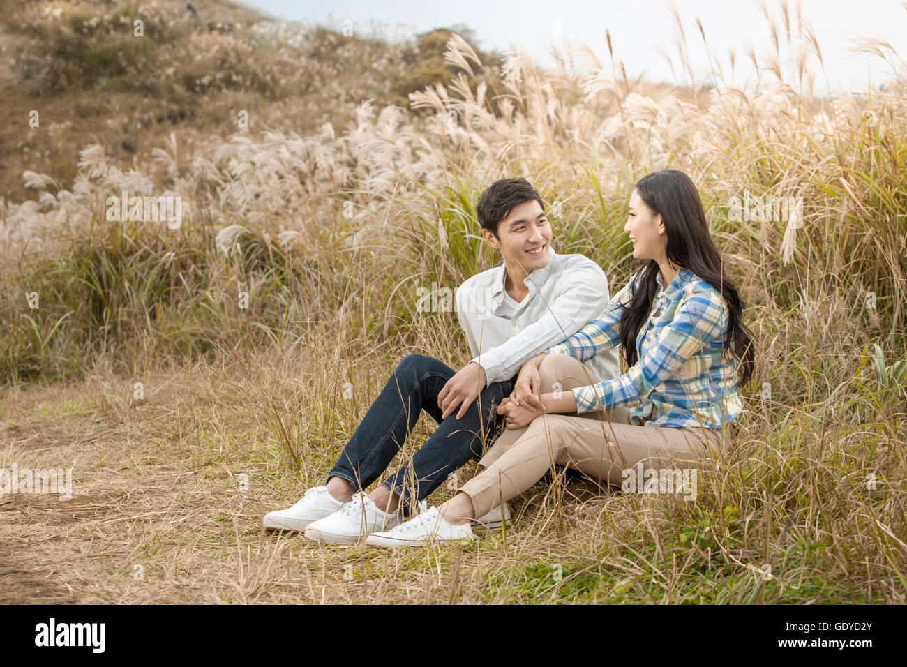 Smiling young couple sitting side by side face to face in silver grass field Stock Photo
