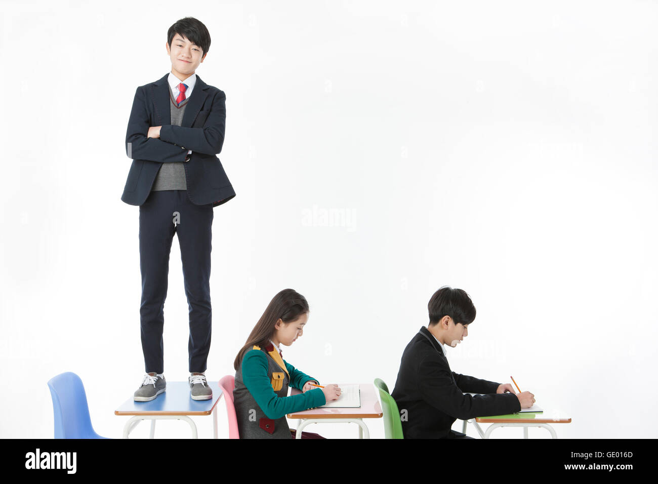 One middle school boy standing on desk with his arms folded and two students studying Stock Photo
