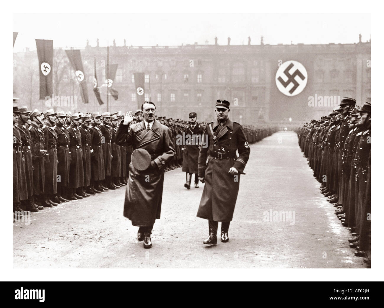 1940's Adolf Hitler in leather trench coat salutes lines of troops with swastika flags in background Stock Photo