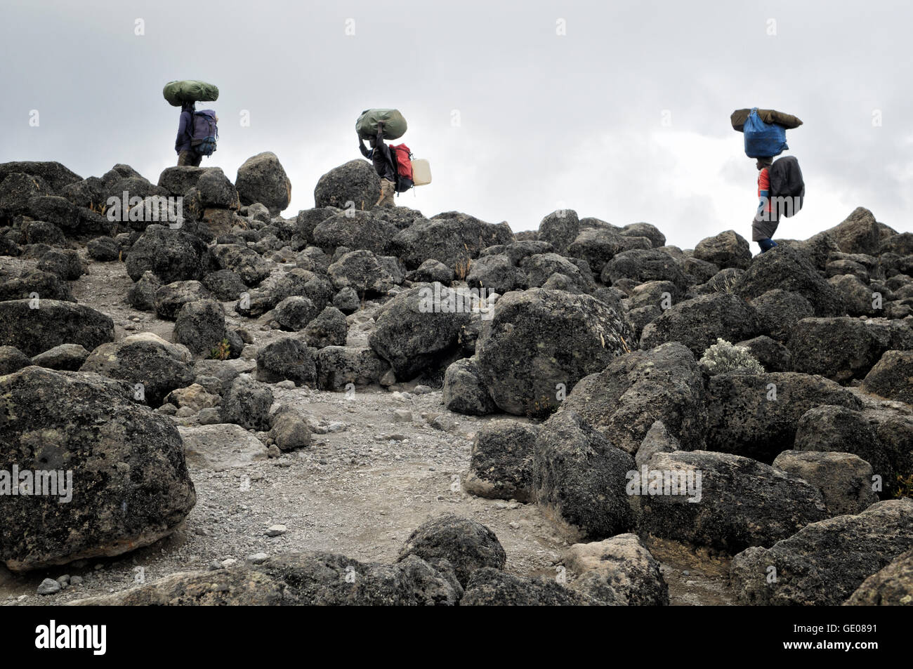 Three porters carrying big bags on their head and big volcanic rocks, Mount Kilimanjaro National Park, Tanzania Stock Photo