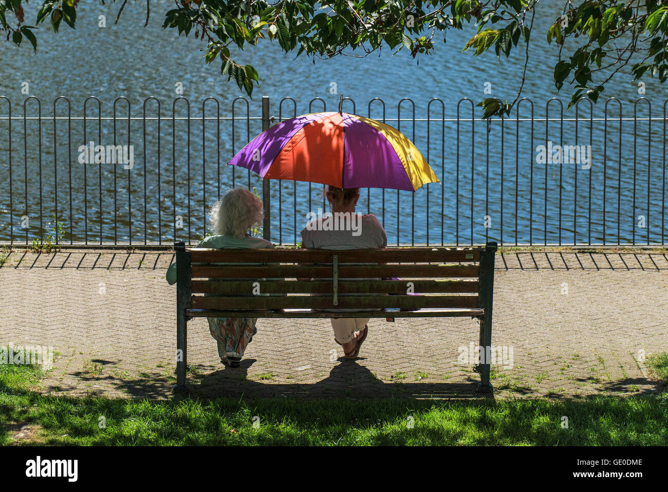 Two women relax in the shade under a colourful umbrella during bright summer sunshine. Stock Photo