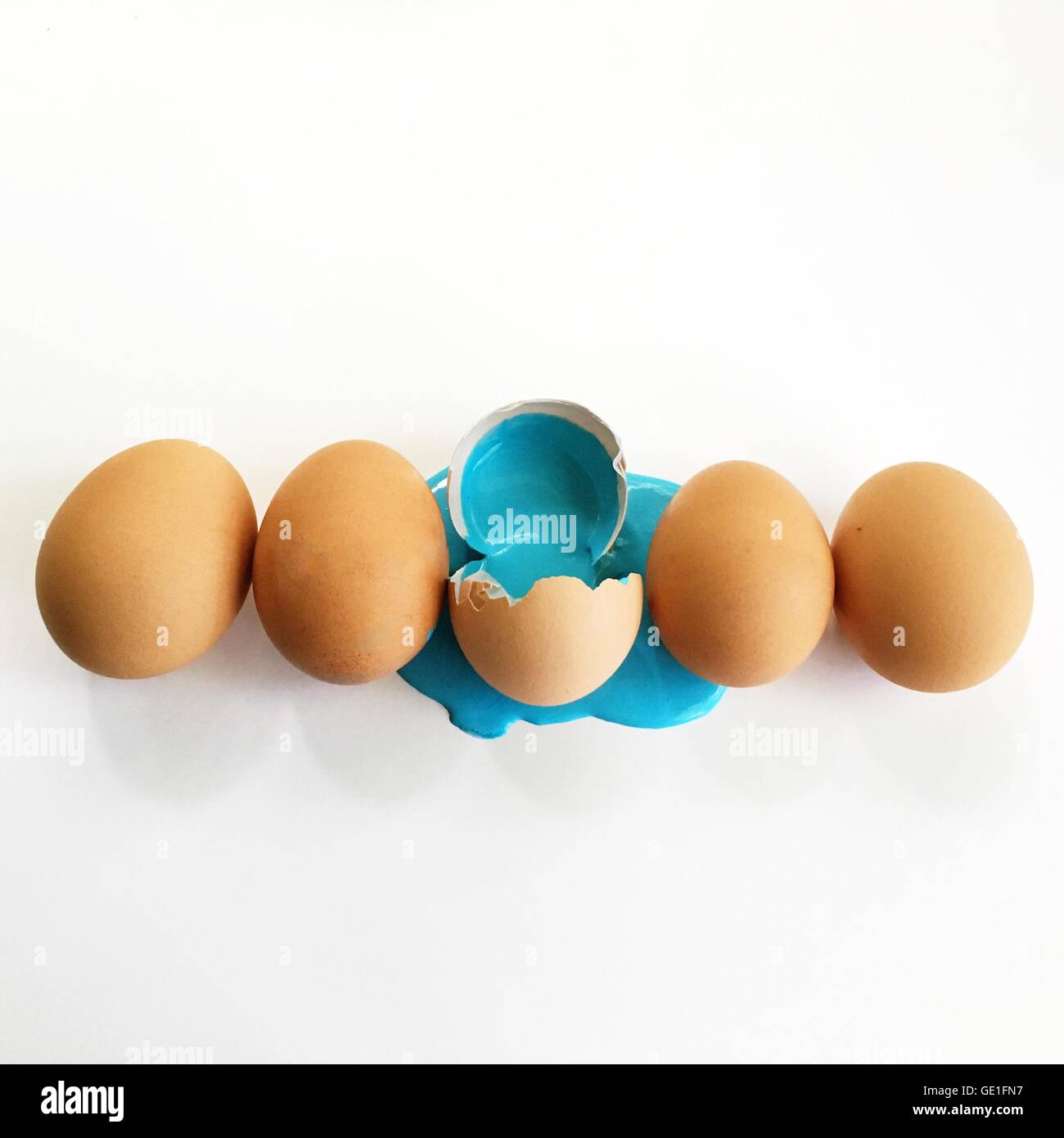 Row of eggs with one cracked open and blue paint spilling out Stock Photo