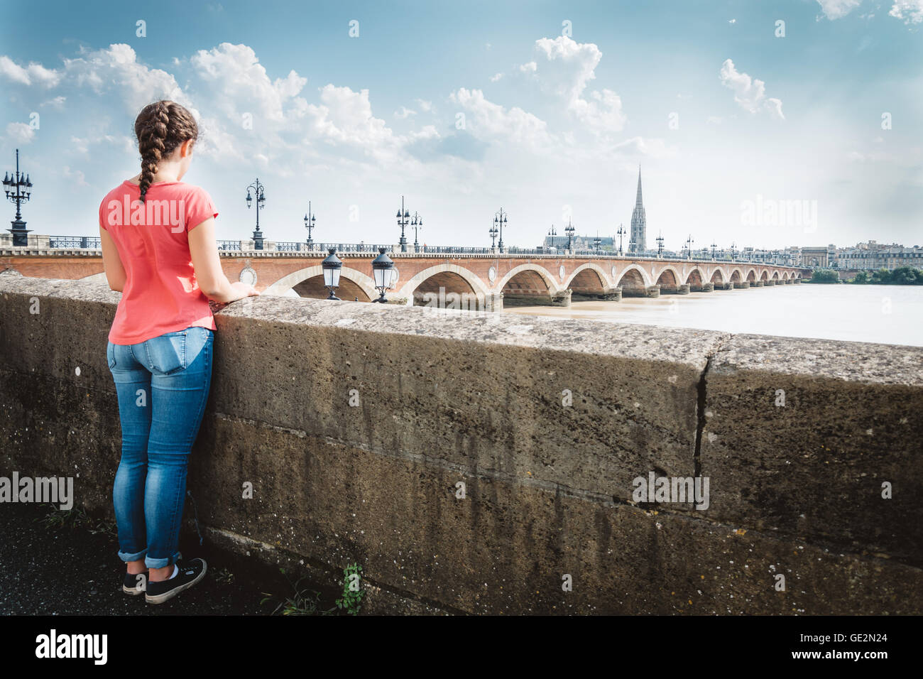 Girl looking at the bridge in Bordeaux a sunny day. Casual wearing a pink tshirt and blue jeans. Rear view Stock Photo