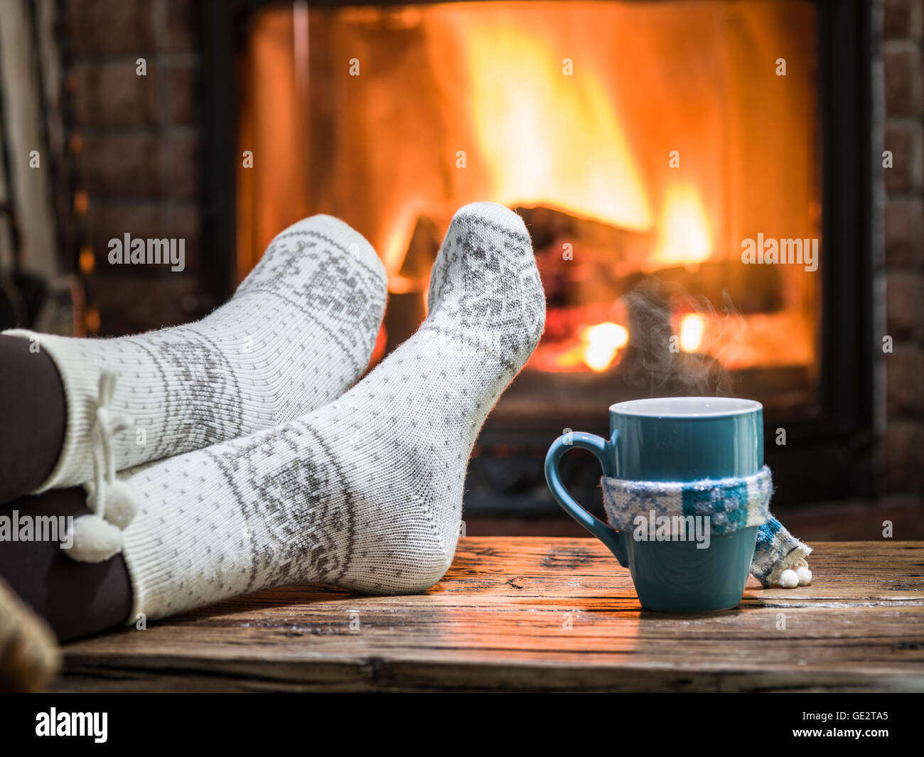 Warming and relaxing near fireplace. Woman feet near the cup of hot drink in front of fire. Stock Photo