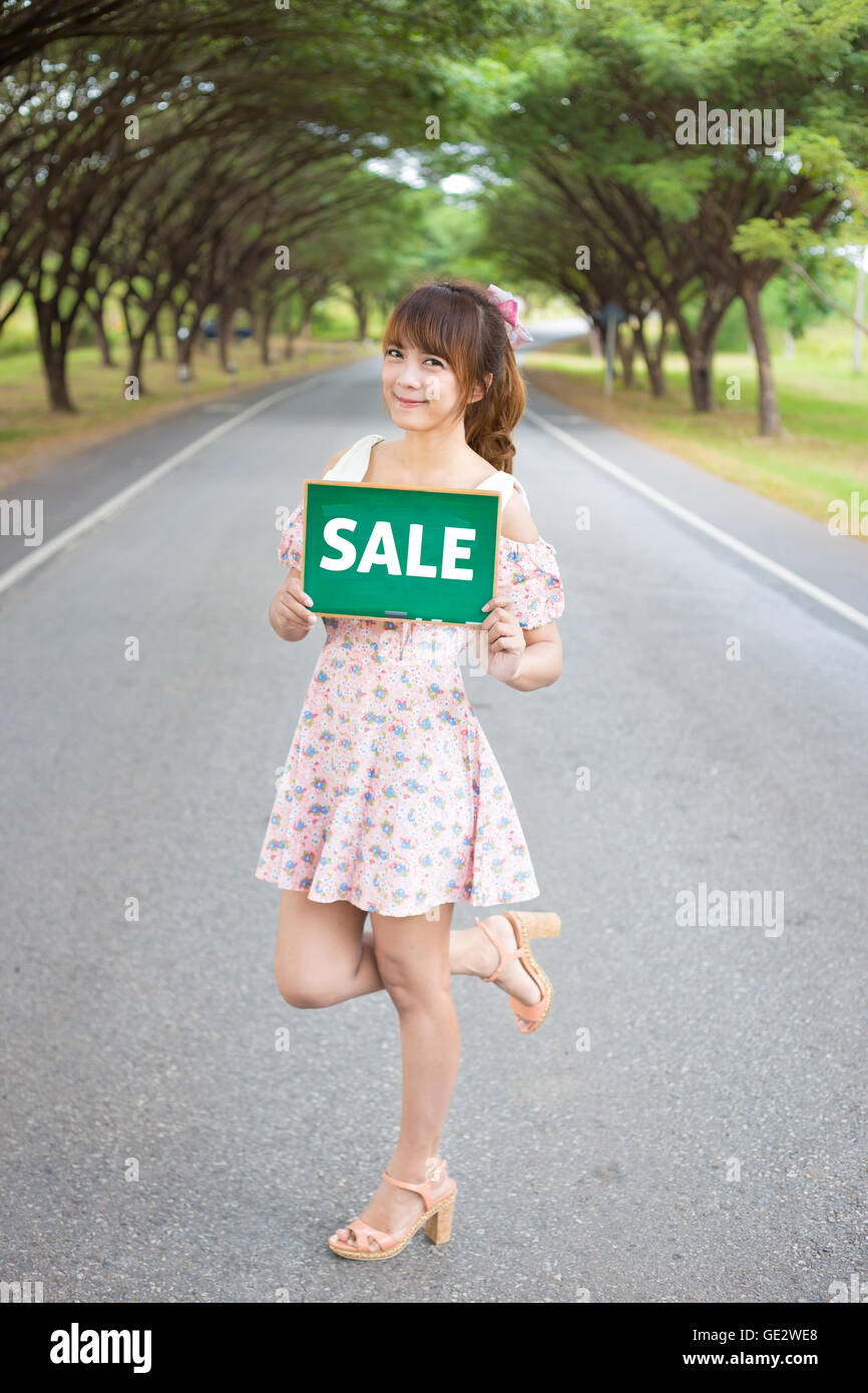 Cute woman hand holding green board sign with text ' sale ' on road and tree, Smiling female model. Stock Photo