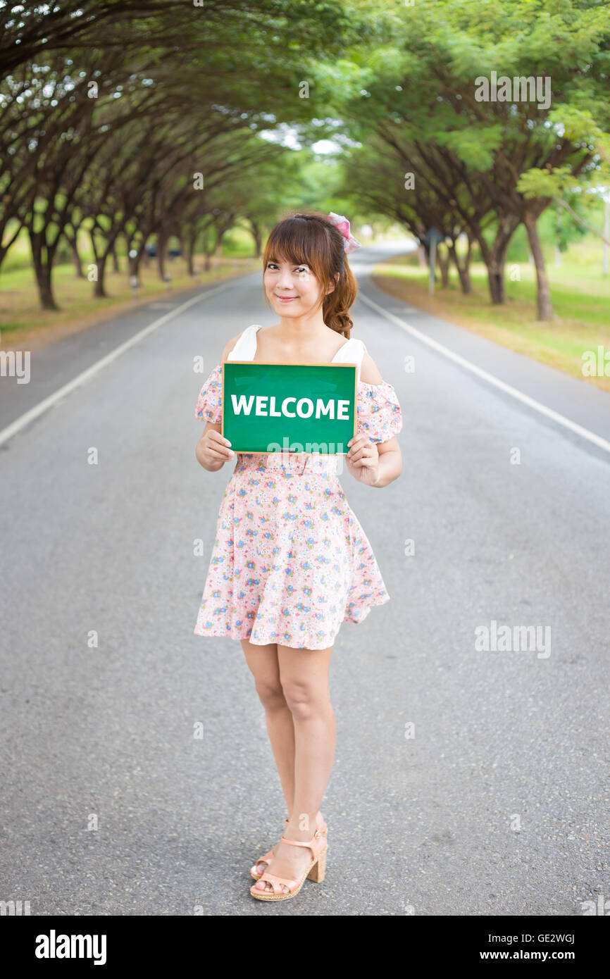 Cute woman hand holding green board sign with text ' welcome ' on road and tree, Smiling female model. Stock Photo