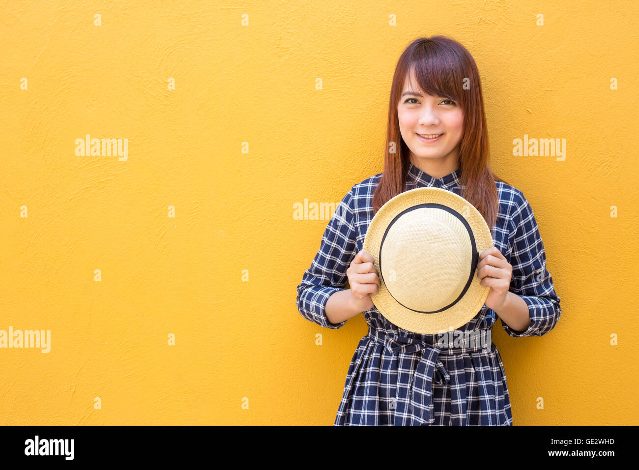 smiling woman wear in dress hand holding hat on yellow cement wall background Stock Photo
