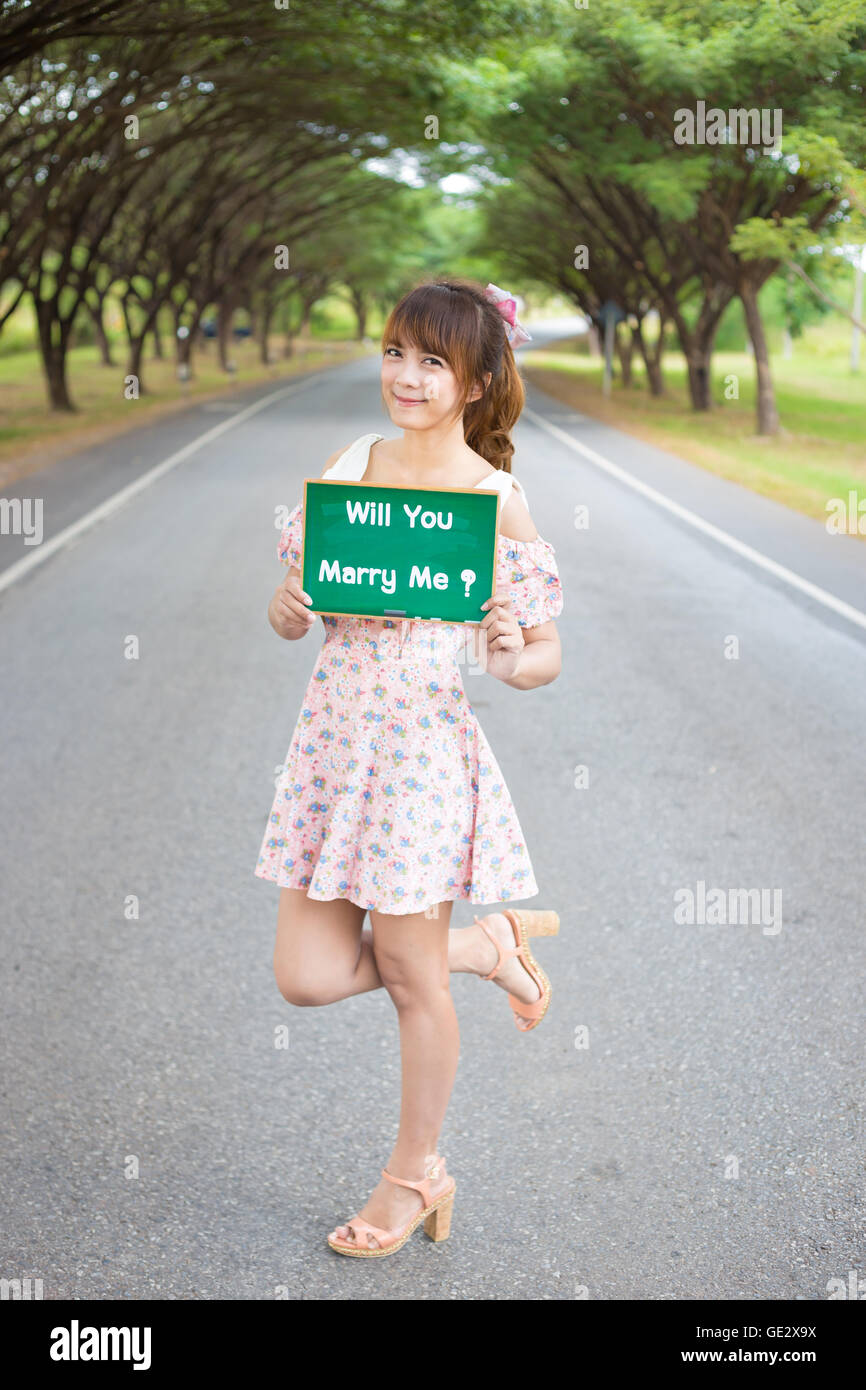 Cute woman hand holding green board sign with text ' will you marry me'on road and tree, Smiling female model. Stock Photo