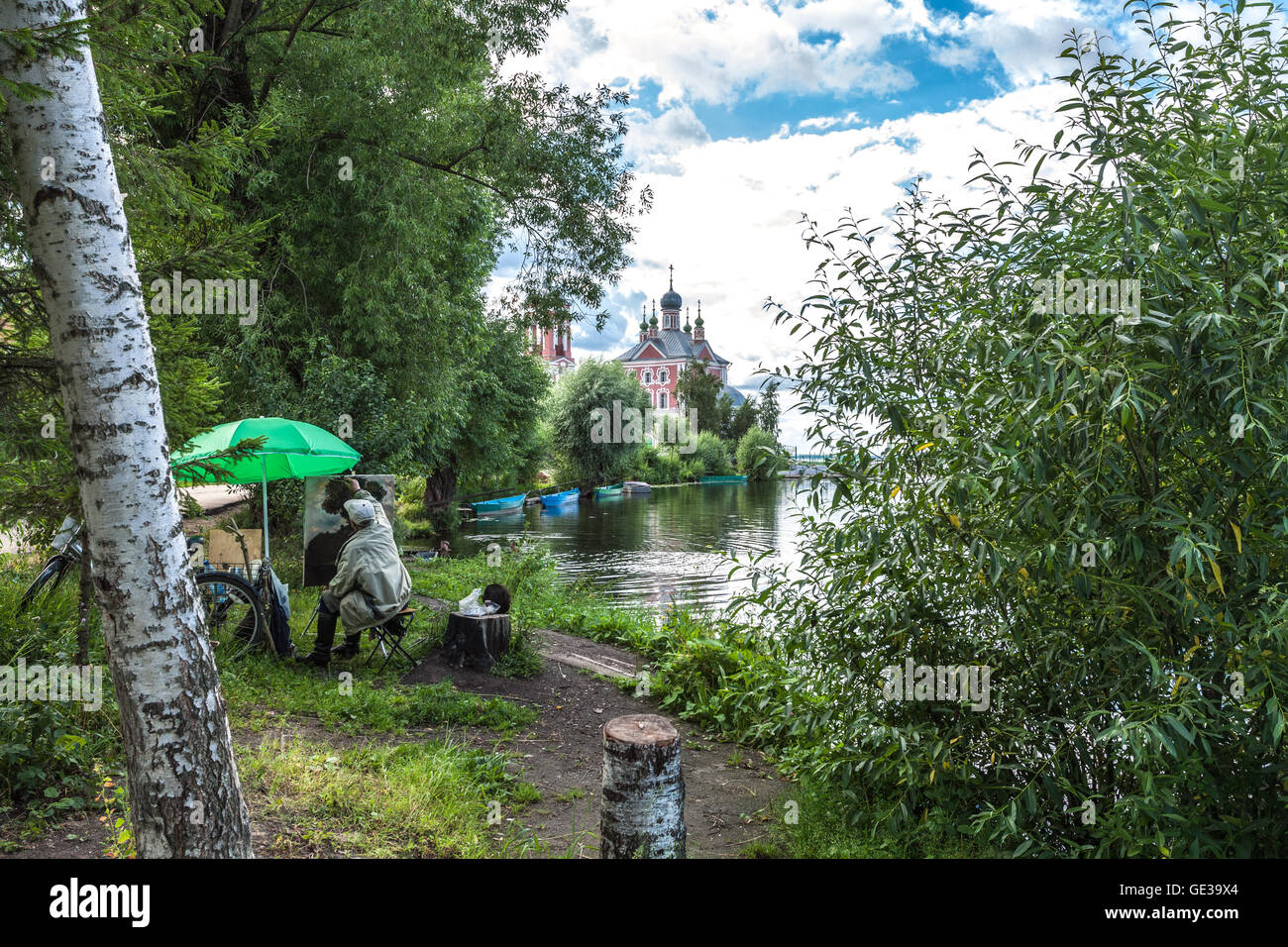 The painter in leather boots and with a black cat on hemp draws an imperceptible tree on other river bank though directly before Stock Photo