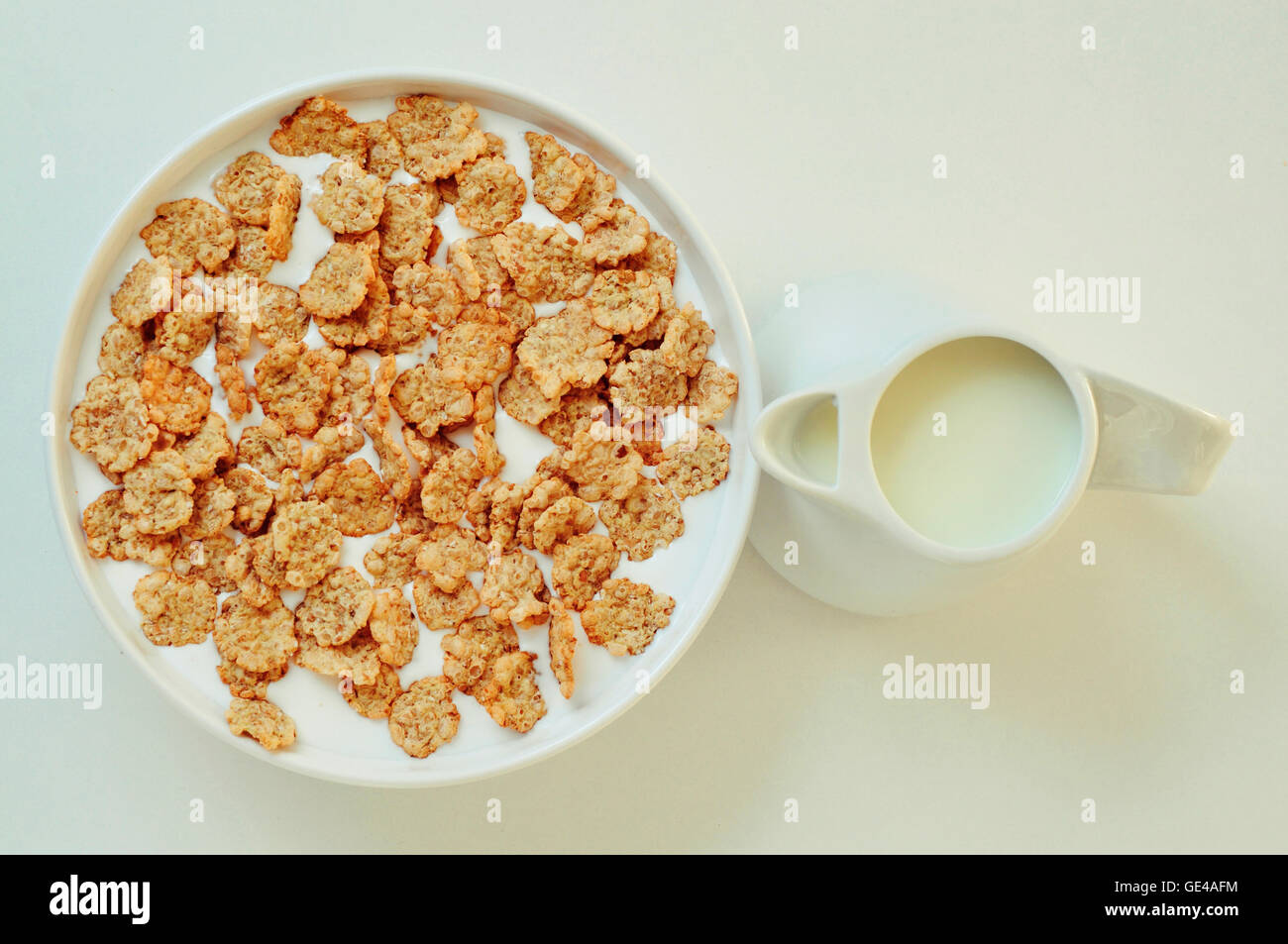 high-angle shot of a white bowl with breakfast cereals soaked in milk and a ceramic milk pot on a white table Stock Photo