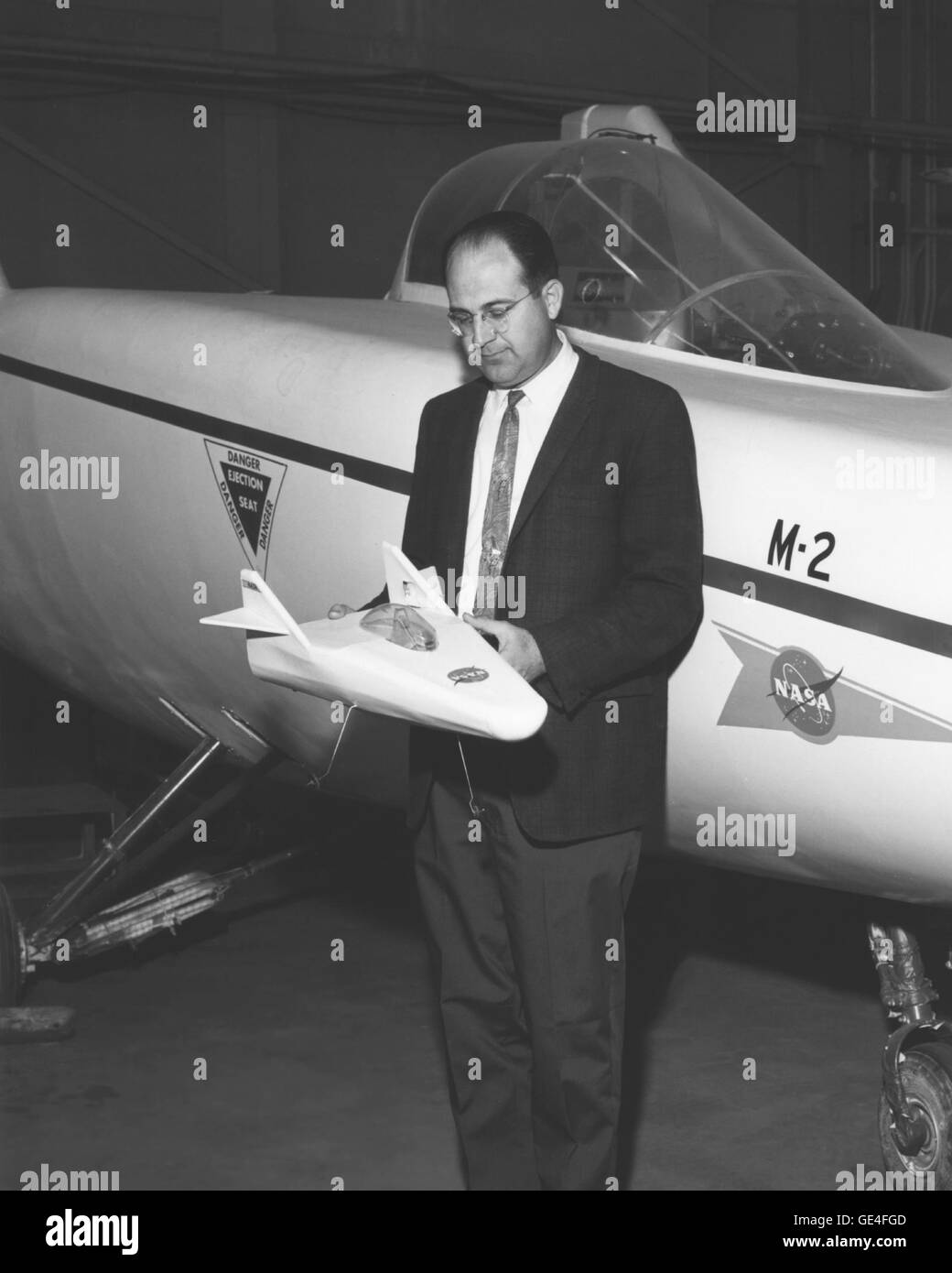 Engineer Dale Reed holds a model of the M2-F1 Lifting Body aircraft with the full scale version directly behind him. In support of the M2 lifting body program in the early 1960s, Dale Reed had built a number of small lifting body shapes and drop tested them from a radio controlled mothership.   Image # : E-16475 Date: March 6, 1967 Stock Photo