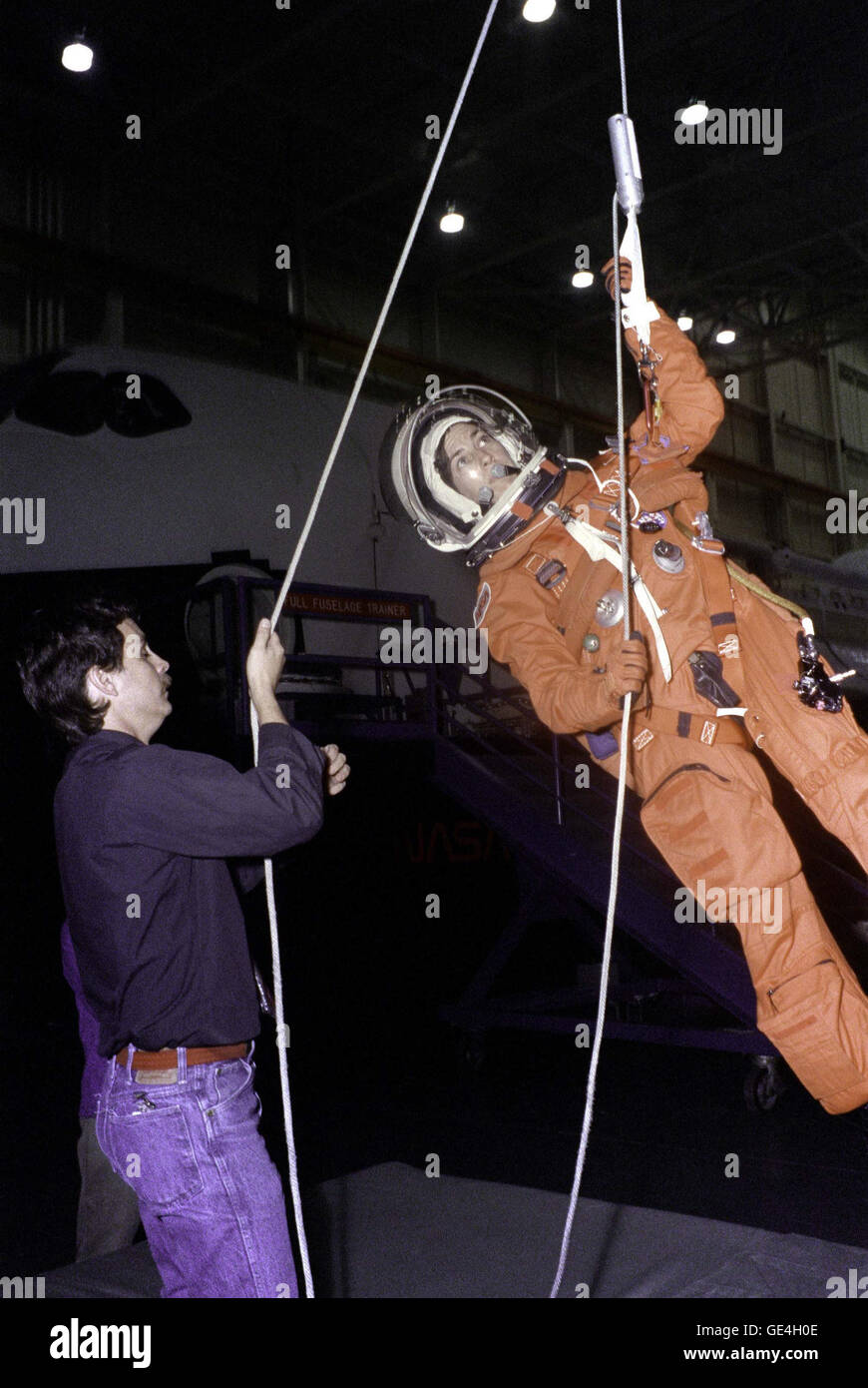 (December 1, 1992) Mission Specialist Ellen Ochoa, wearing a Launch and Entry Suit (LES) and Launch and Entry Helmet (LEH), simulates an emergency egress procedure at JSC's Mockup and Integration Laboratory (MAIL). Having exited the crew compartment trainer (CCT) a shuttle mockup, through an overhead aft flight deck window; Ochoa lowers herself to the ground using the sky-genie. Training instructor Kenneth D. Trujillo assumes the role of a crewmate assisting from a position on the ground. The sky-genie is carried on all Space Shuttle flights for emergency egress purposes.   Image # : S92-51716 Stock Photo