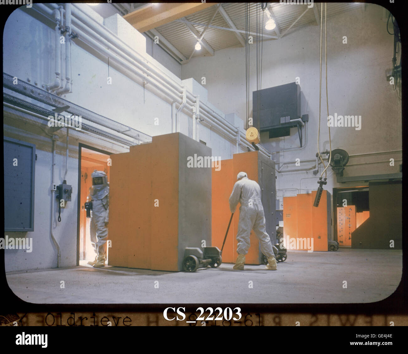 A technician emerges from the rear of a hot laboratory cell in full protective gear carrying a &quot;cutie pie&quot; radiation detector. Another technician wheels open the massive sixty-inch-thick concrete door plug. Stock Photo