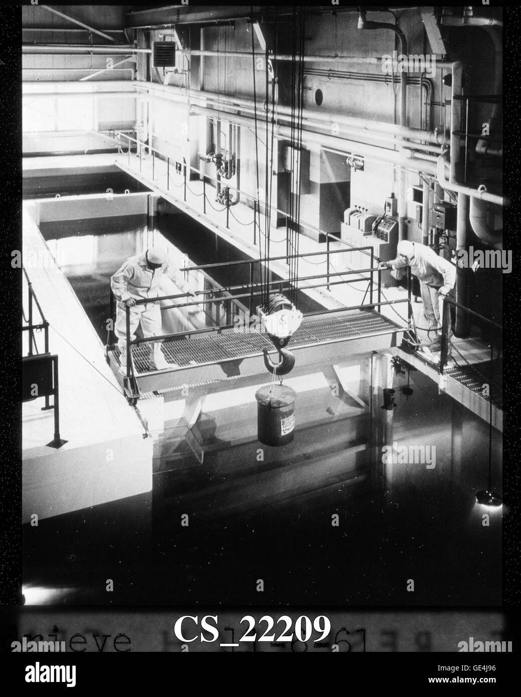 Two Plum Brook employees use an overhead crane to lift a lead cask of low- level radioactive waste from Canal F. This was the first canal outside of the containment vessel. Canals G and H are visible behind the man standing on the bridge. The bridge was movable so technicians could continually work above the objects as the moved through the canal system. The canal connected to the hot laboratory, which was adjacent to the south side of the reactor building. Radioactive materials were moved under water with vehicles, or remotely controlled cranes, between heavily shielded walls in the hot handl Stock Photo