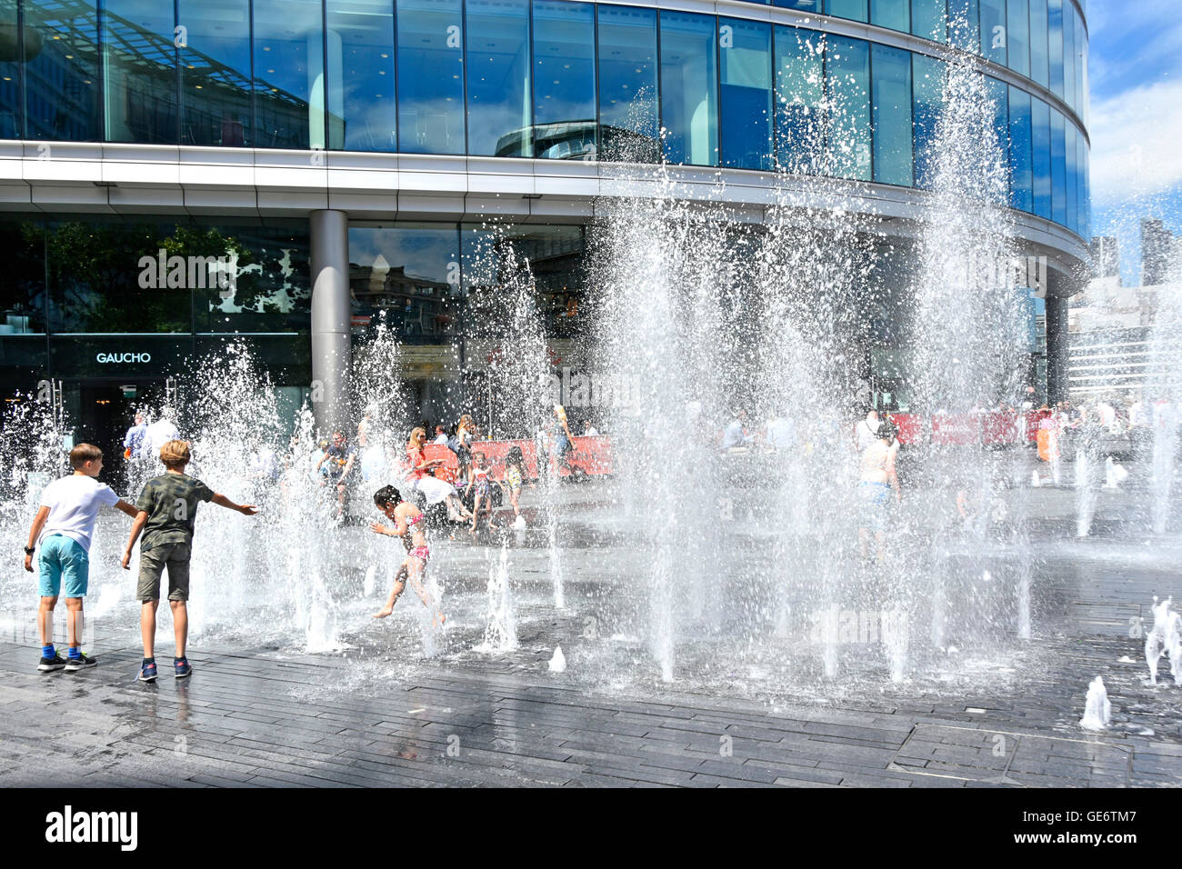Water jets rising above block paving at random timings during hot London UK summer weather as office workers take lunch break & kids play in fountains Stock Photo