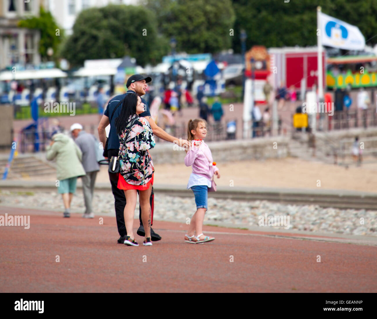 A young family walking down the coastal promenade at Llandudno in North Wales in summer with a young girl looking towards the kite she is holding in the air, Wales, UK Stock Photo