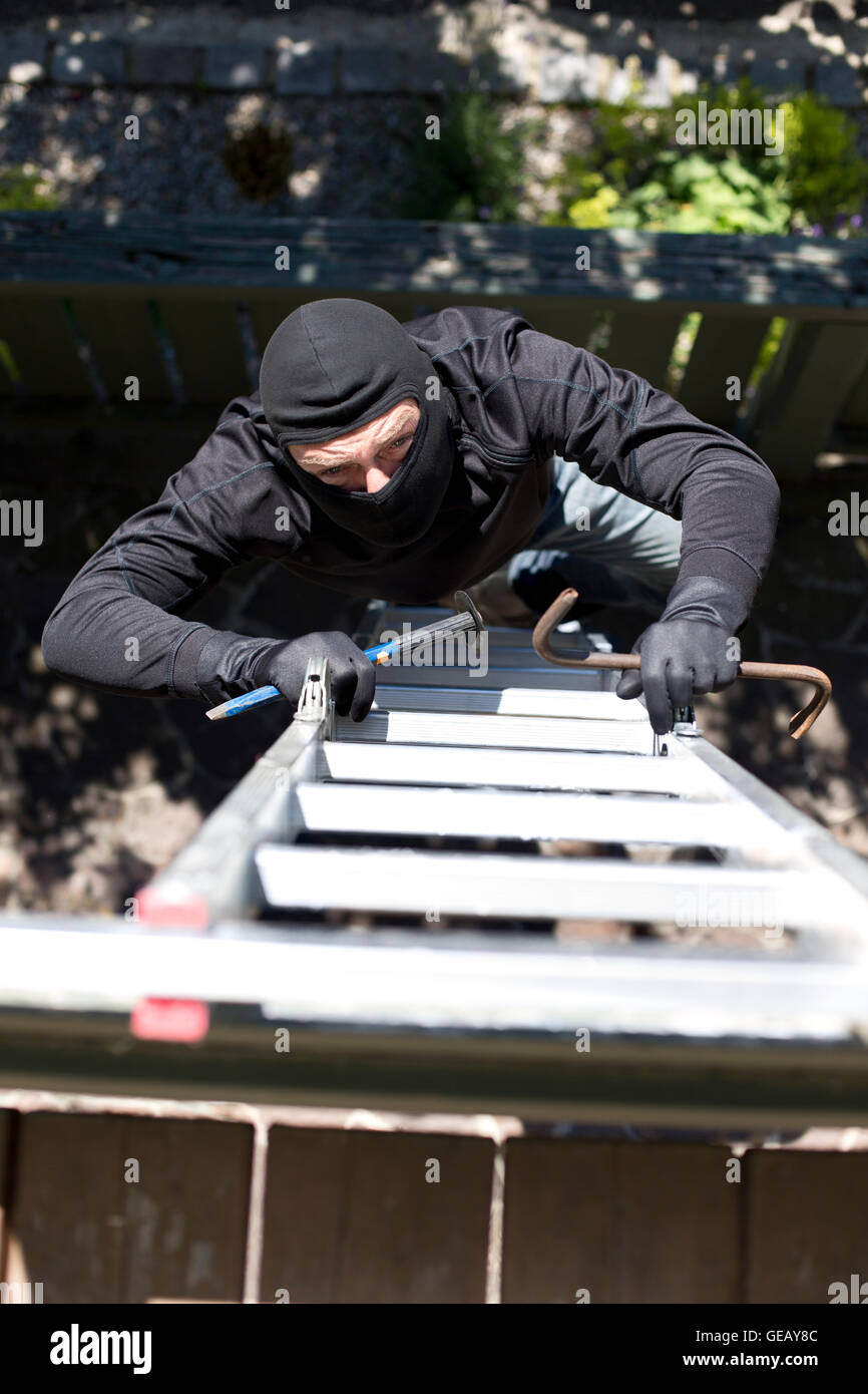 Burglar with crowbar climbing up ladder at house wall Stock Photo