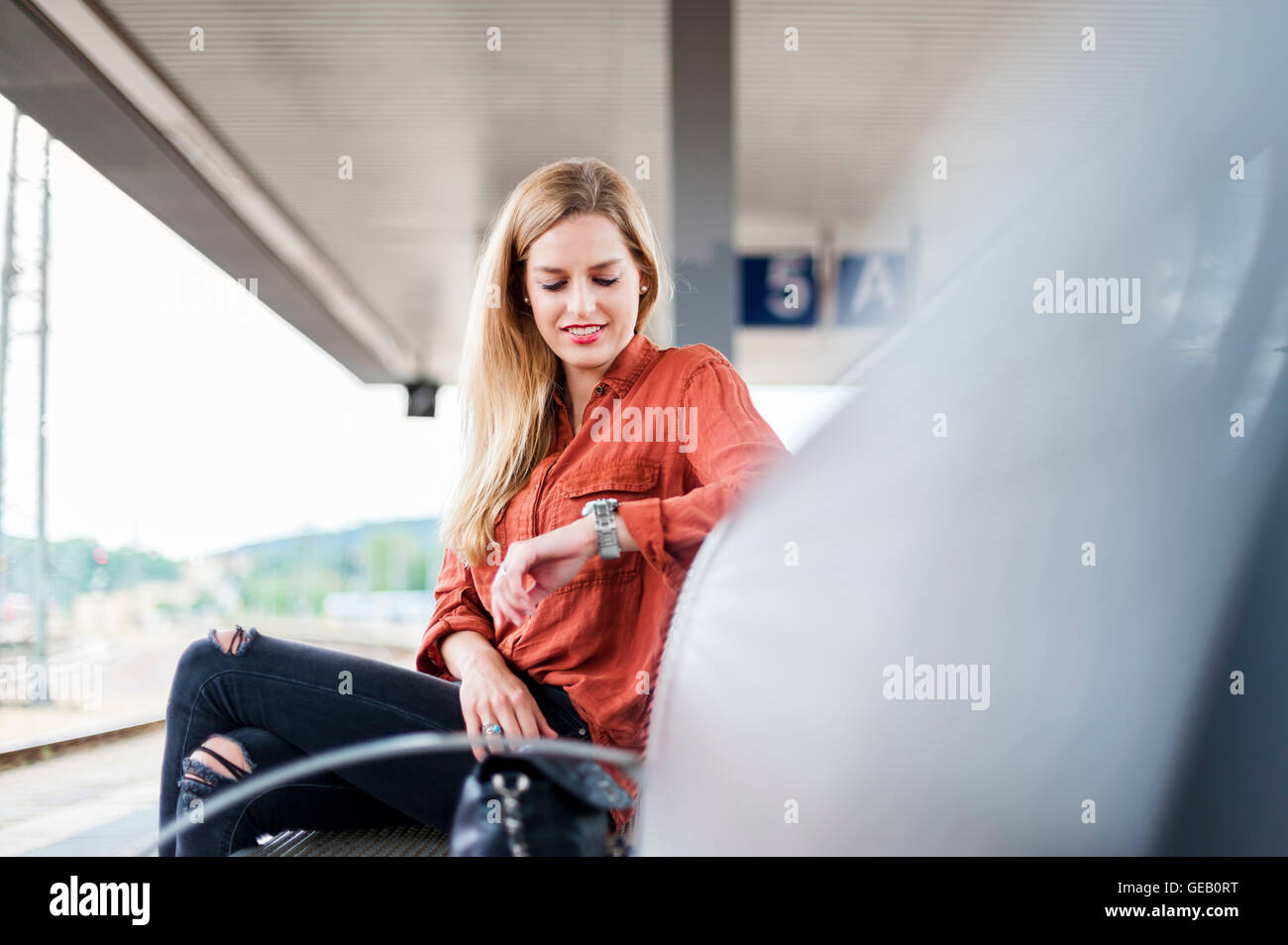 Smiling young woman sitting on bench at platform checking the time Stock Photo