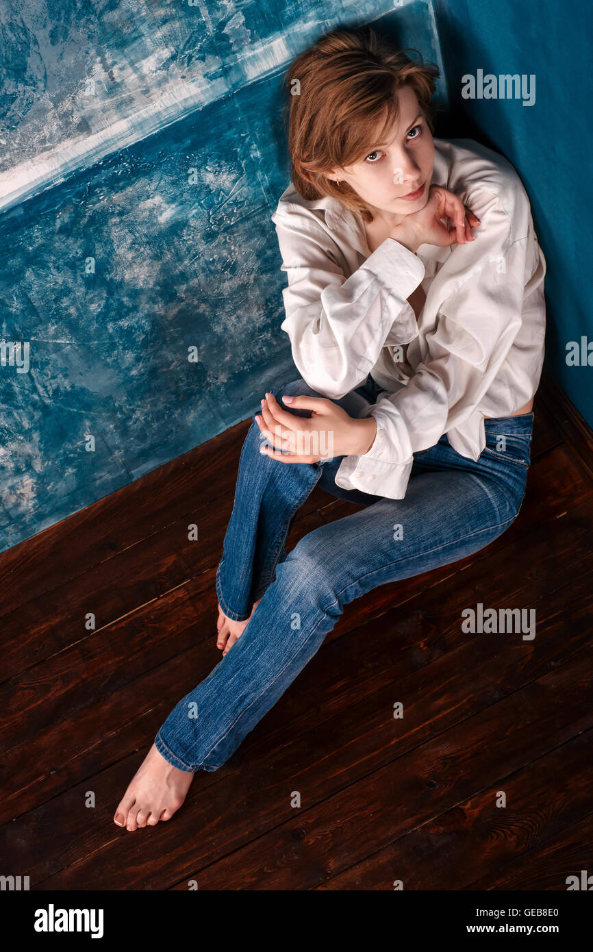 Lonely woman in casual white shirt and blue jeans sitting on the floor in the corner of blue abandoned room. Top view Stock Photo