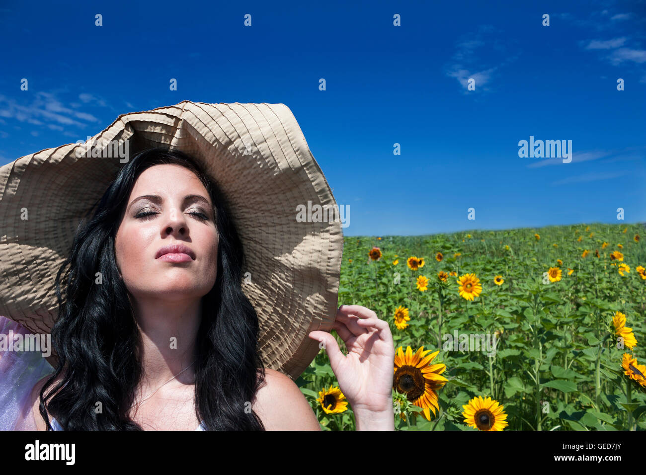 Pretty young brunette woman wearing large hat in a sunflower field ...