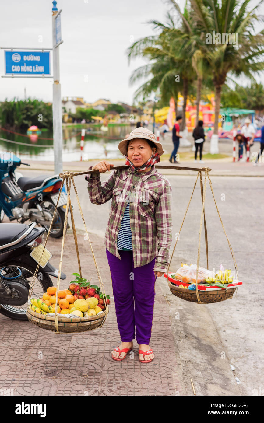 Hoi An, Vietnam - February 16, 2016: Asian seller carrying fresh fruit in bowls on her shoulders in the street in Hoi An, Vietnam. Rambutan, mango, banana and mandarin. Stock Photo