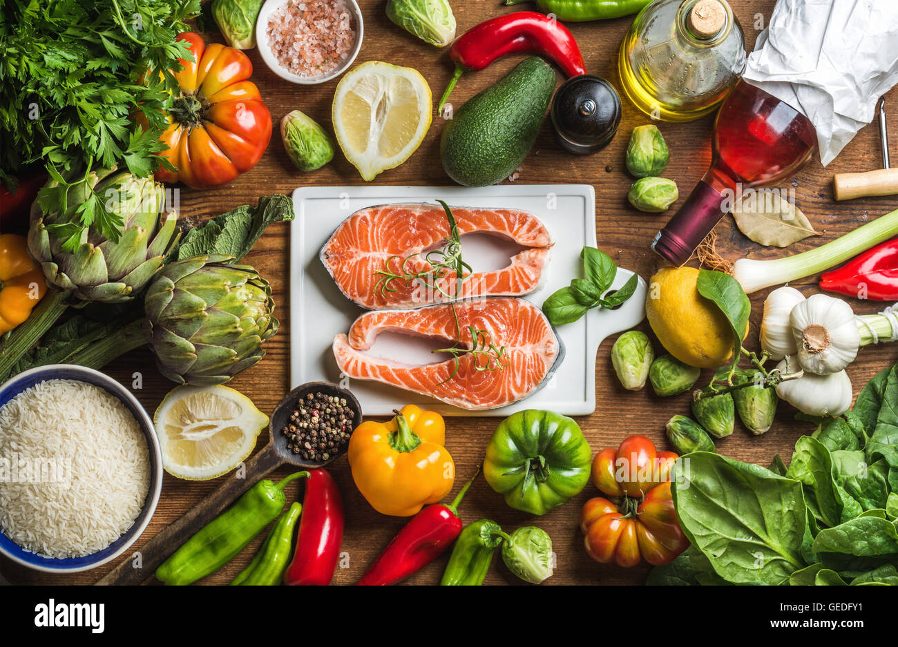 Dinner cooking ingredients. Raw uncooked salmon fish with vegetables, rice, herbs, lemon, artichokes, spices and bottle of rose wine on white ceramic board over rustic wooden background, top view Stock Photo