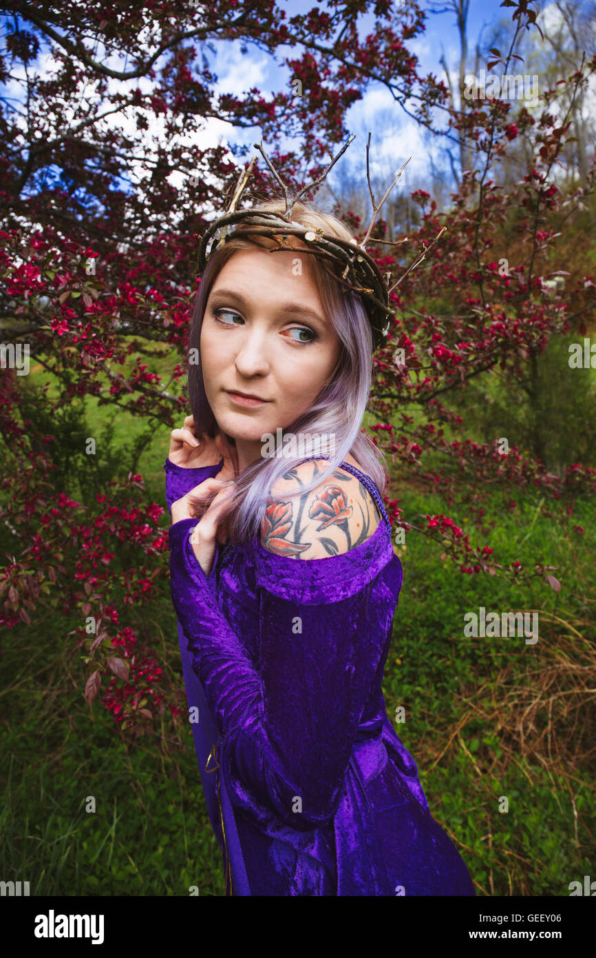 Young woman dressed in a purple velvet gown and twig crown with flowering crab apples in the background Stock Photo