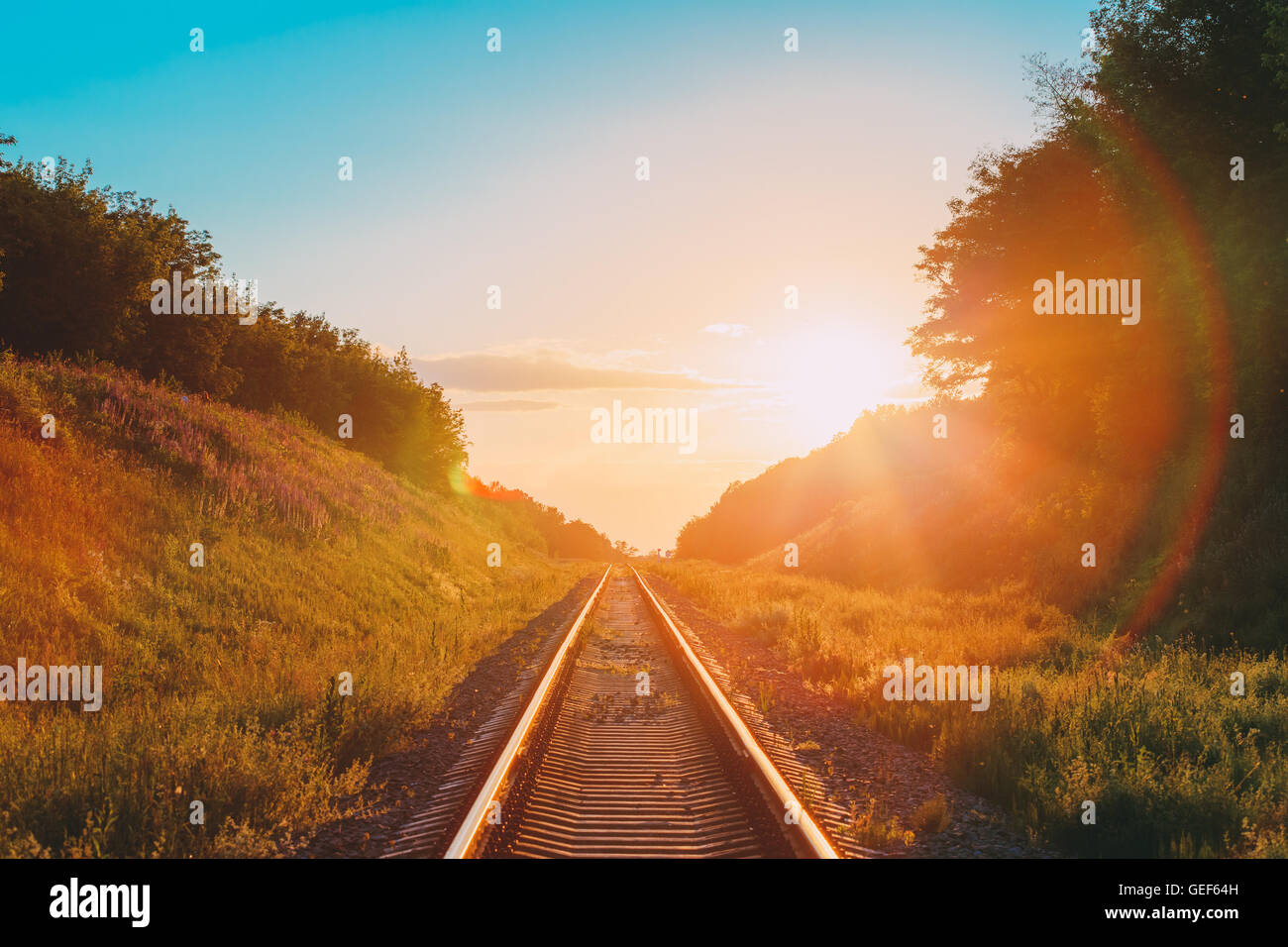 The Scenic Landscape With Railway Going Straight Ahead Through Summer Hilly Meadow To Sunset Or Sunrise In Sunlight. Lense Flare Stock Photo
