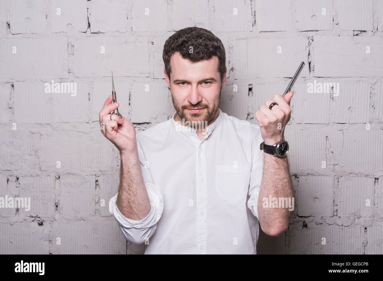 Young hairdresser, wearing in white shirt, posing with scissors and comb, on grey brick wall background Stock Photo