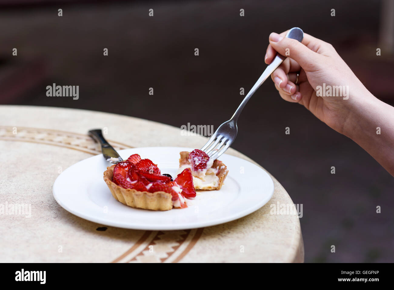 Photo of female hands cutting a fruit cake with strawberries, mint and blackcurrant, on a white plate on a restaurant table. Stock Photo